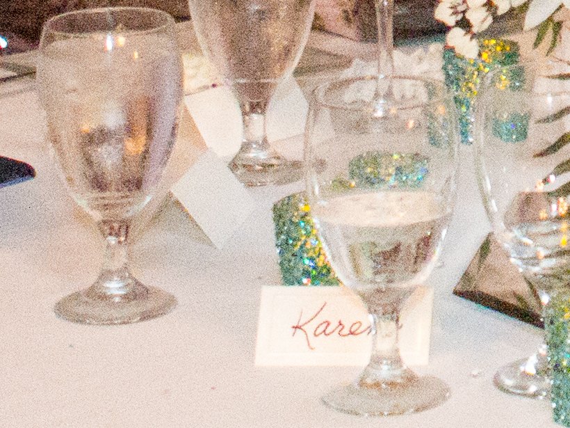 Two water glasses, a handwritten name card reading Karen and a glittering candle holder on a white tablecloth at a wedding reception, speckled all over with grain in a photograph taken at ISO 12800