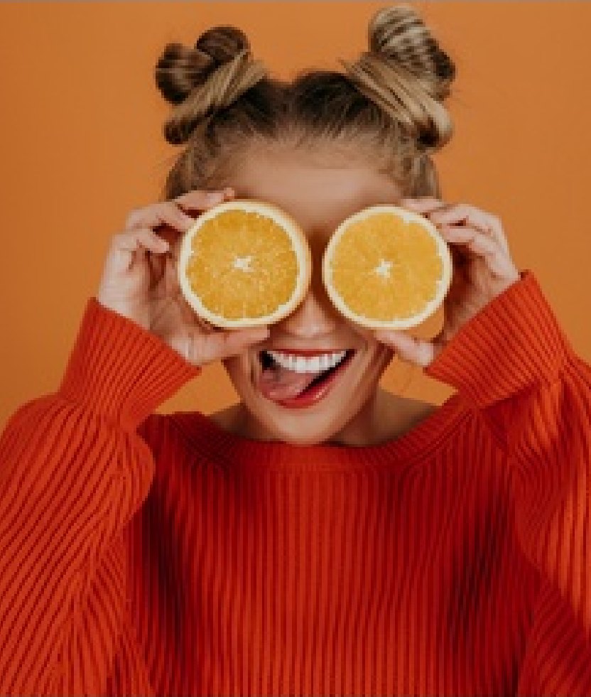 A young woman with her hair in two buns, in an orange sweater, holding orange halves over her eyes and sticking out her tongue in front of an orange wall