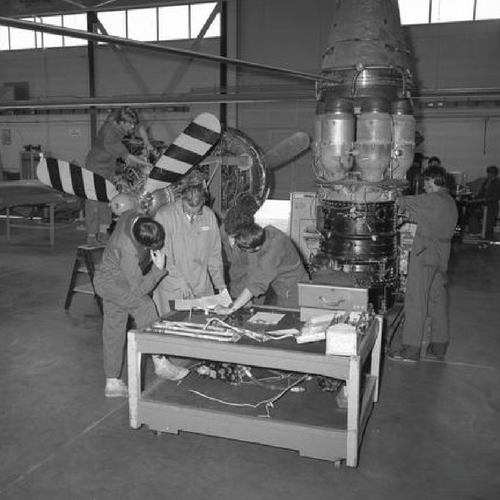Technicians in coveralls and a man in a lab coat studying a plan at a workbench in an aircraft hangar, beside a propeller and an engine on a stand, in a small black-and-white photograph