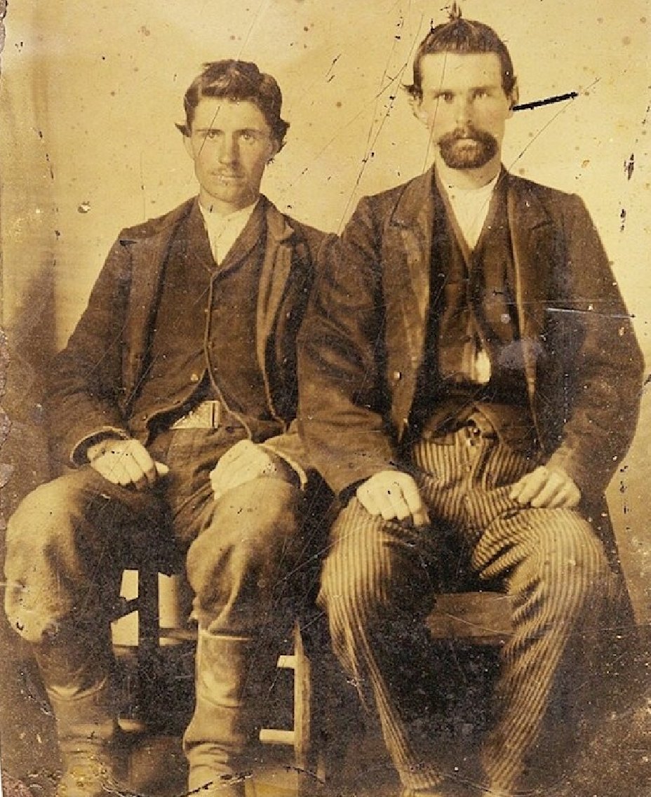 Two young men in dark jackets seated side by side, one with a beard, in a scratched and stained sepia tintype