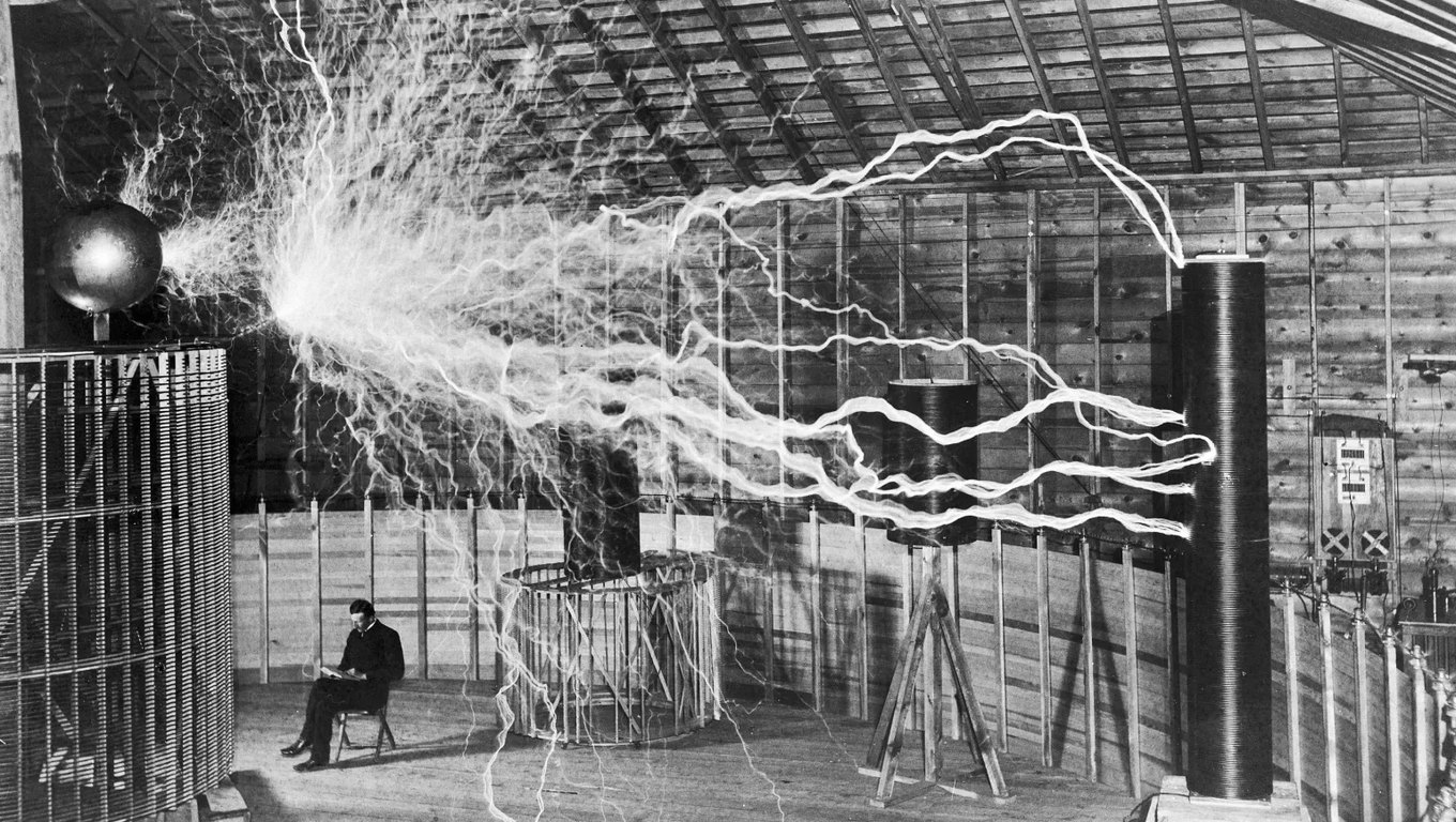 Nikola Tesla reading in his Colorado Springs laboratory beneath arcs of electricity from a giant coil, in a black-and-white photograph from 1899