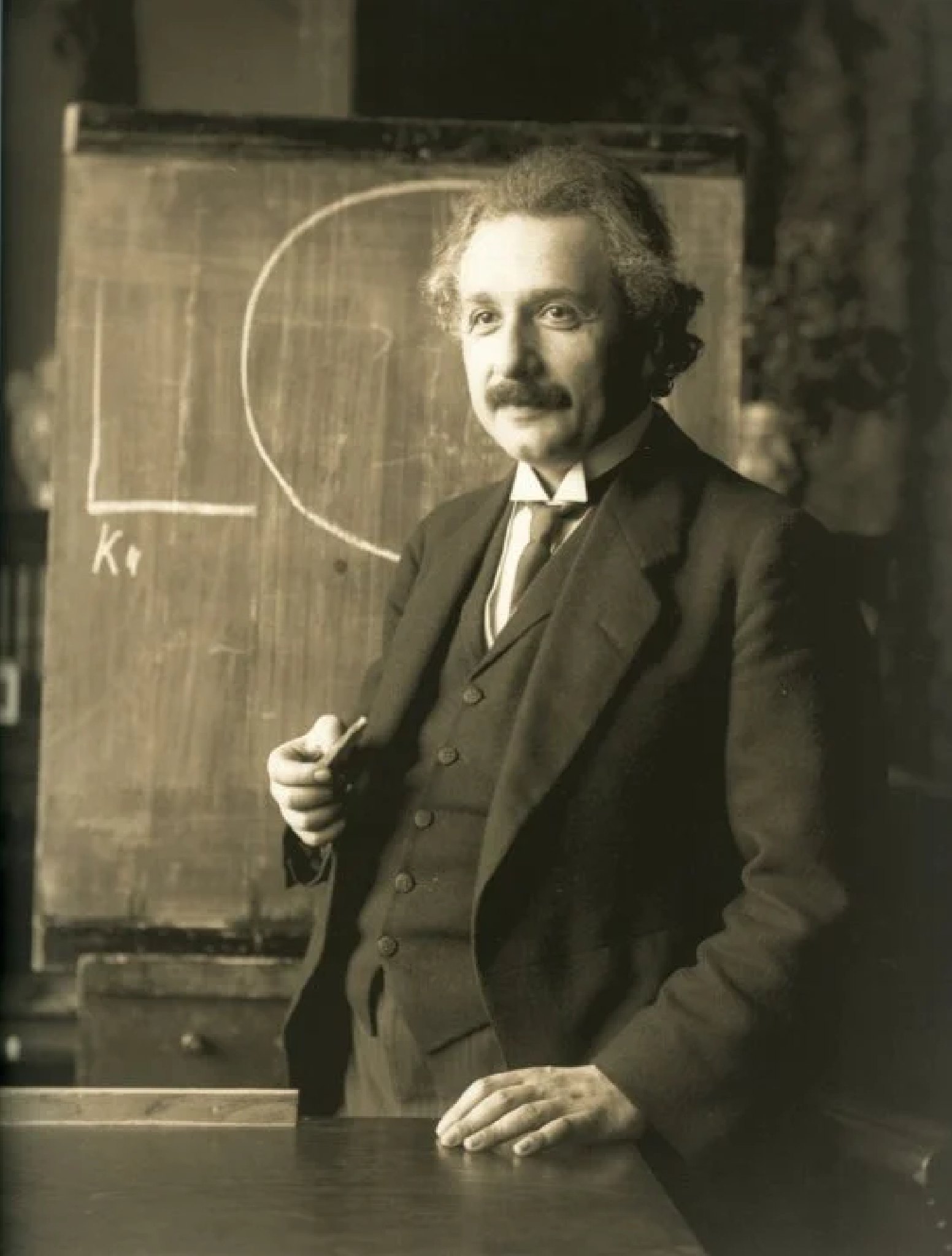 Albert Einstein at a wooden blackboard in a faded sepia photograph, chalk in his right hand