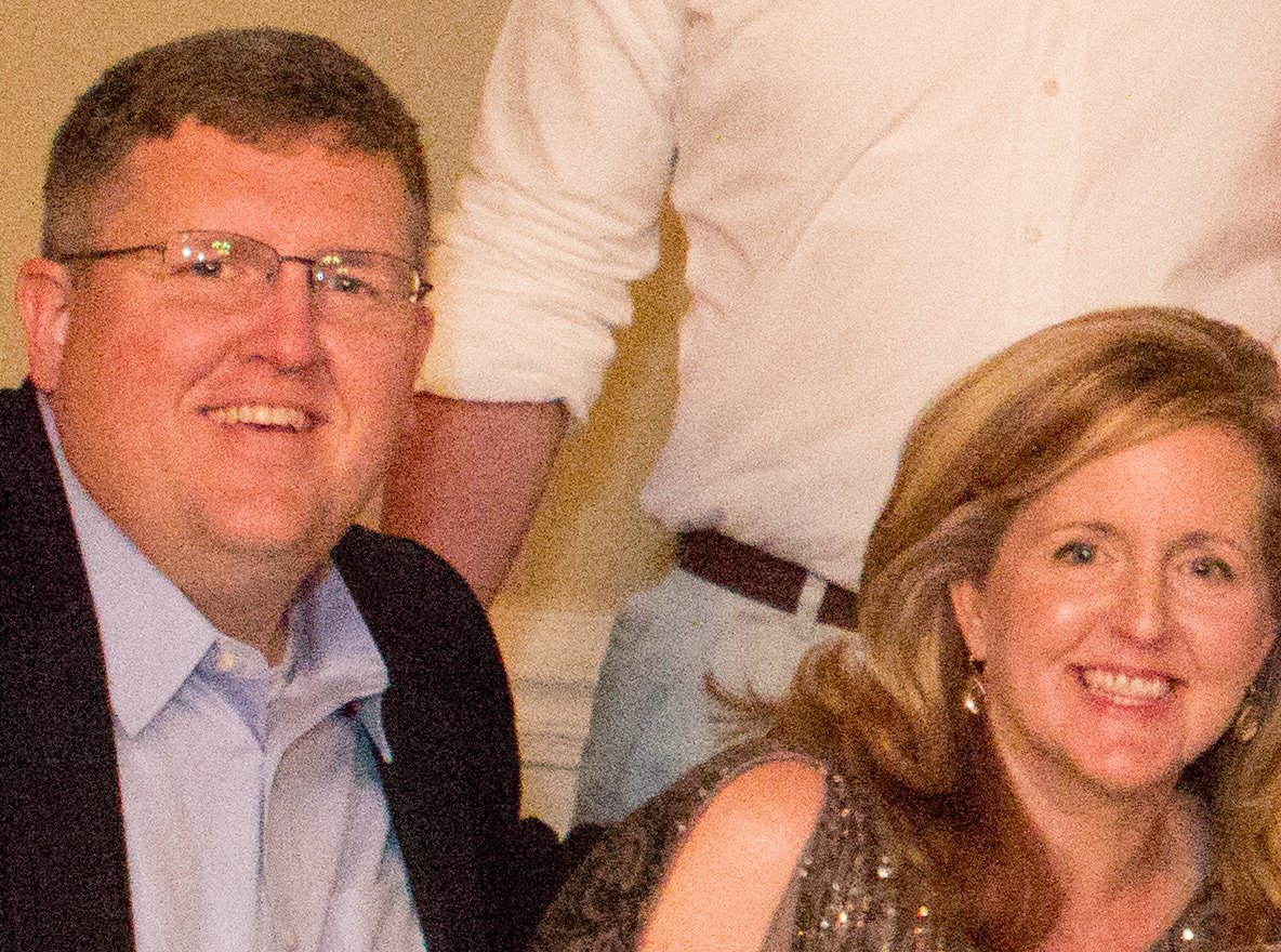The same smiling man in rimless glasses and smiling woman with long fair hair at the reception table, covered in heavy coloured grain from a high ISO